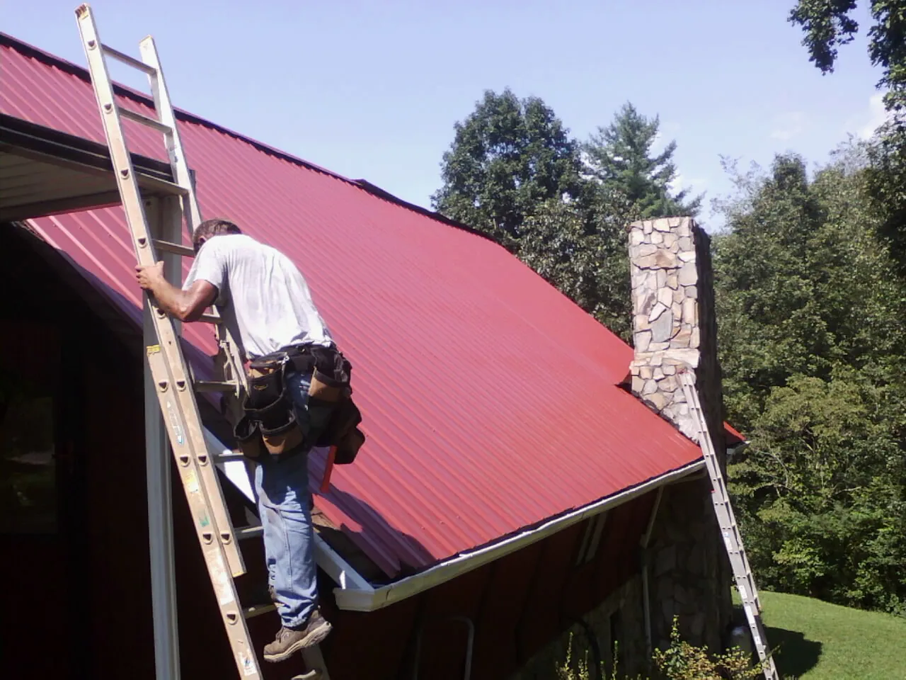 Roofer on a ladder beside a red metal roof and stone chimney — Soffit & Fascia Repair by Roofing Brothers in Tamiment
